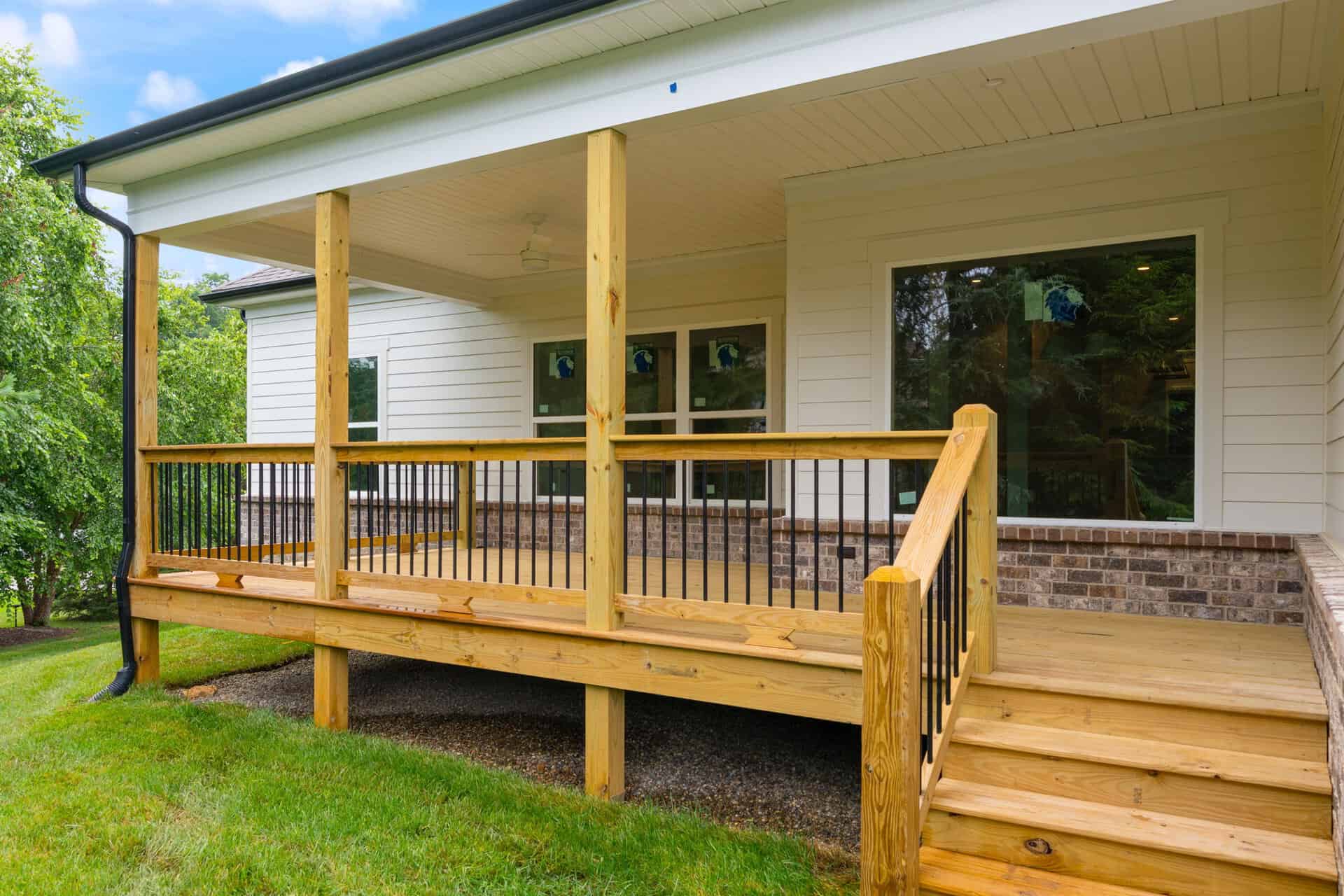 Front porch of a light-colored house with wooden railing, steps, and large windows reflecting green trees in the yard.