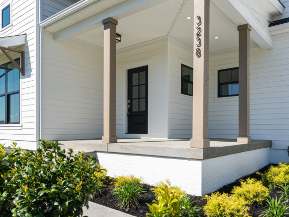 White house with a covered front porch, beige posts, and a dark front door; neatly landscaped shrubs line the walkway.