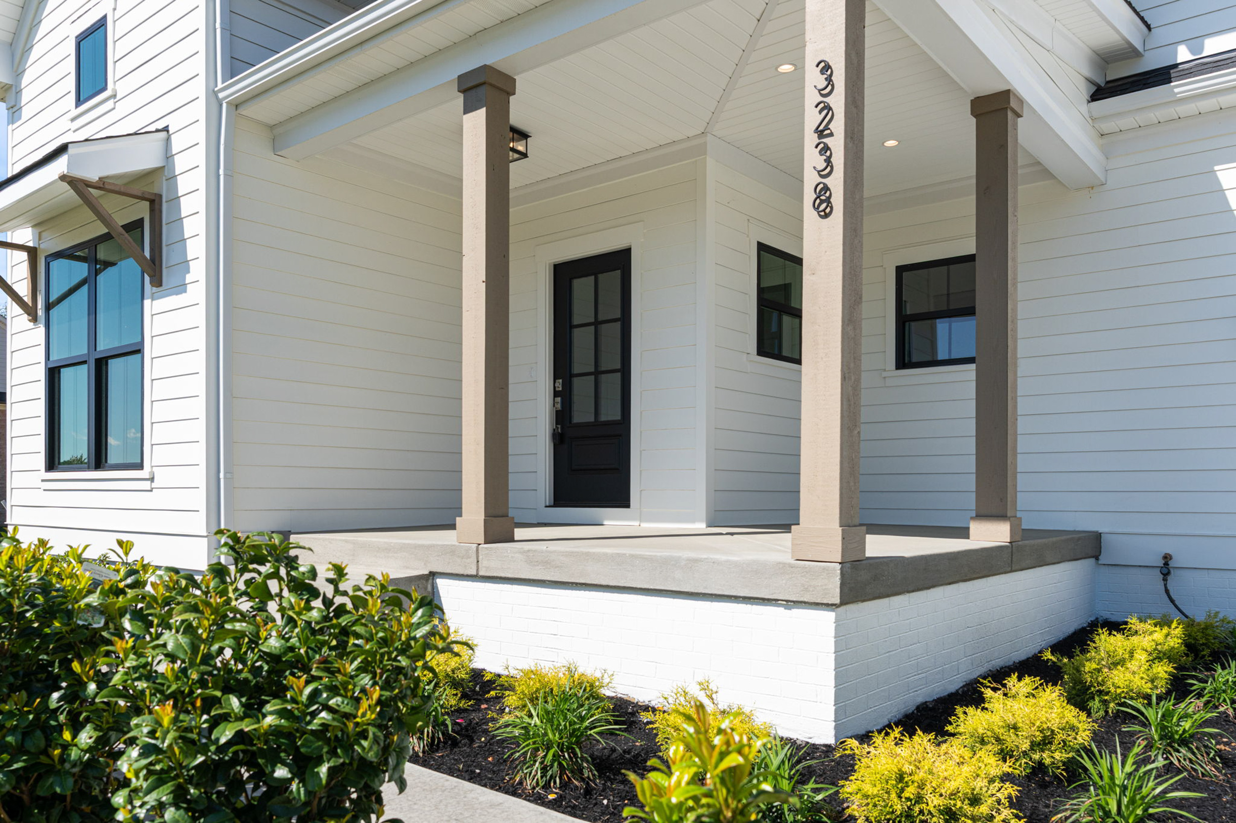 White house with a covered front porch, beige posts, and a dark front door; neatly landscaped shrubs line the walkway.