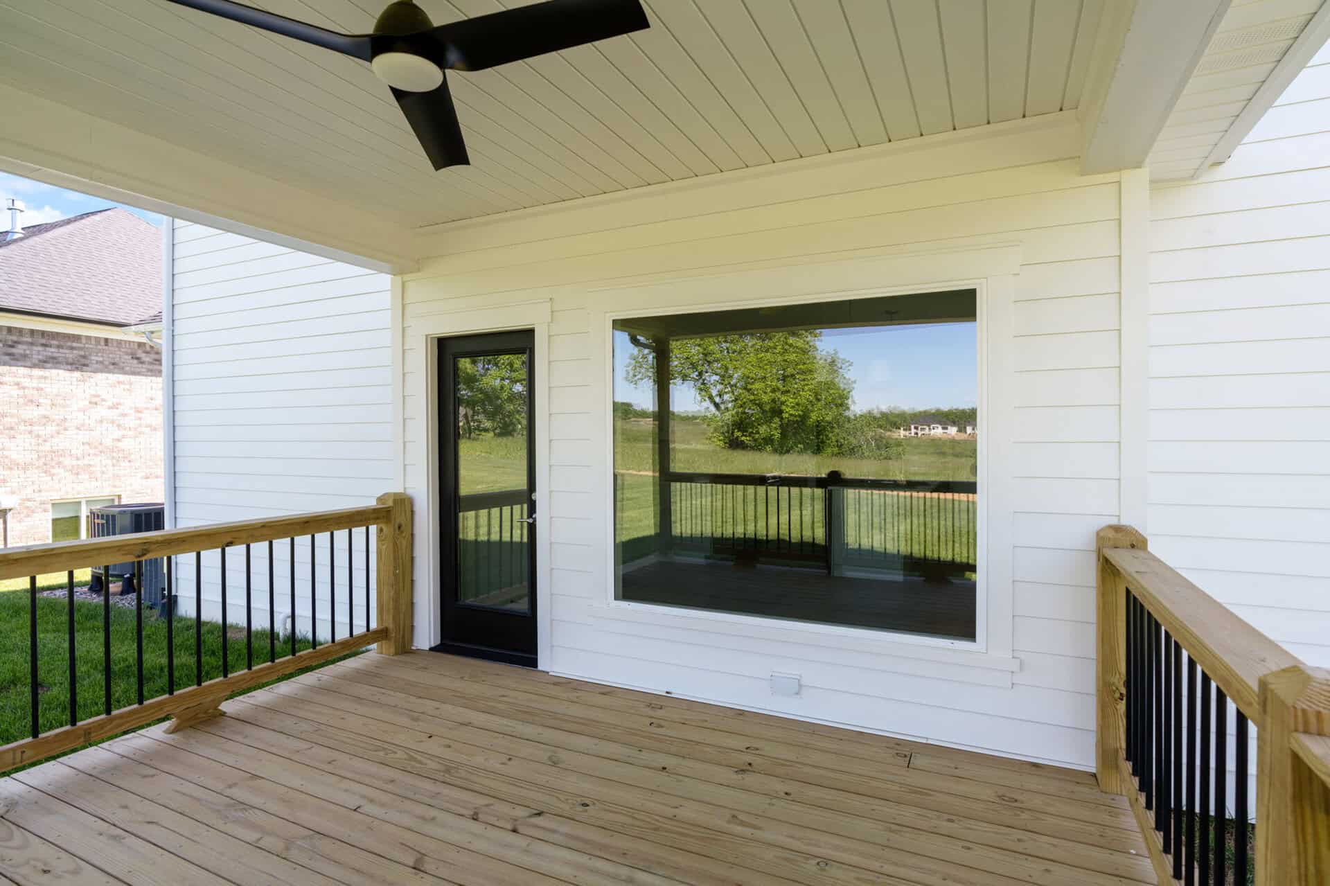 White siding house with a glass door and large window opening onto a wooden deck; a black ceiling fan hangs from the covered porch roof, and green yard is visible beyond.