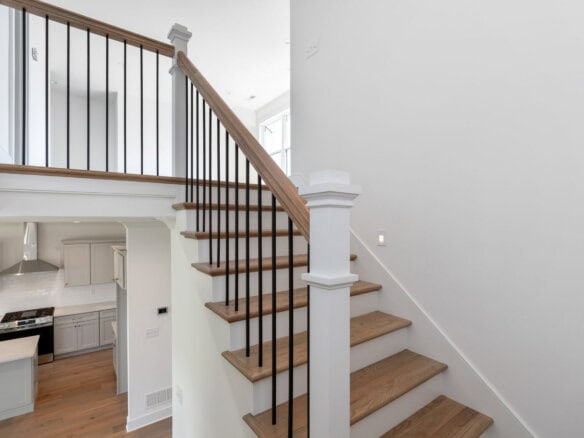Open two-story staircase with wooden steps and black metal balusters beside a white wall, with the kitchen visible below.