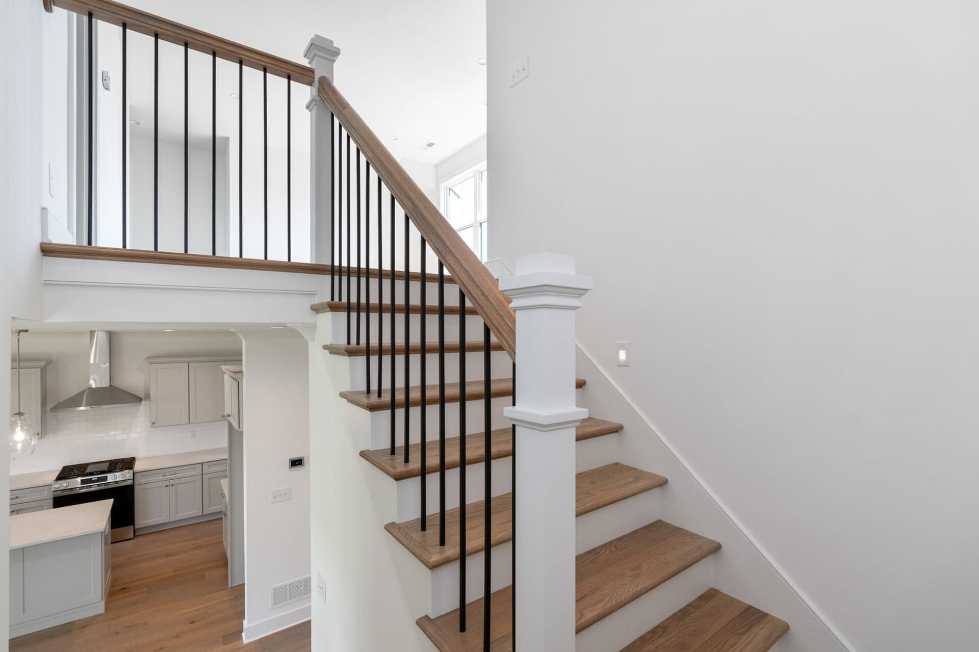 Open two-story staircase with wooden steps and black metal balusters beside a white wall, with the kitchen visible below.