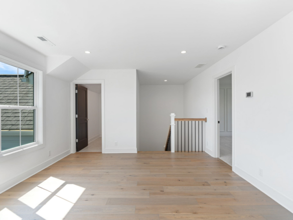 Sunlit upstairs hallway with light hardwood floors, white walls, and a wooden railing by the stairs on the right; large window on the left letting in natural light.