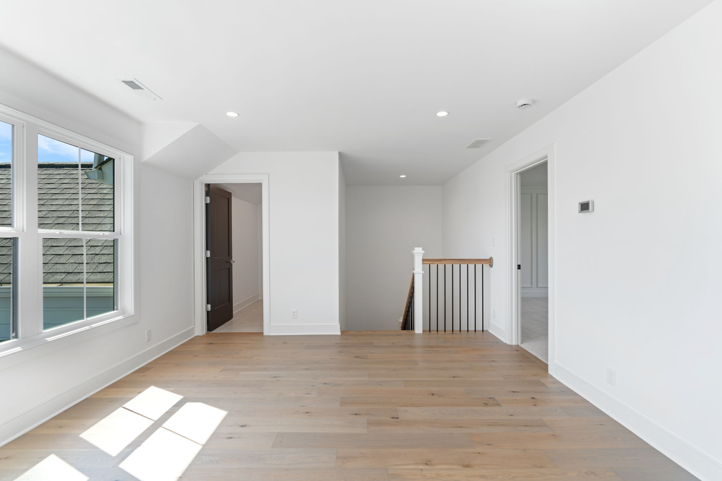 Sunlit upstairs hallway with light hardwood floors, white walls, and a wooden railing by the stairs on the right; large window on the left letting in natural light.
