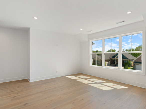 Bright empty living space with a large window and light wood flooring, white walls, and recessed ceiling lights.