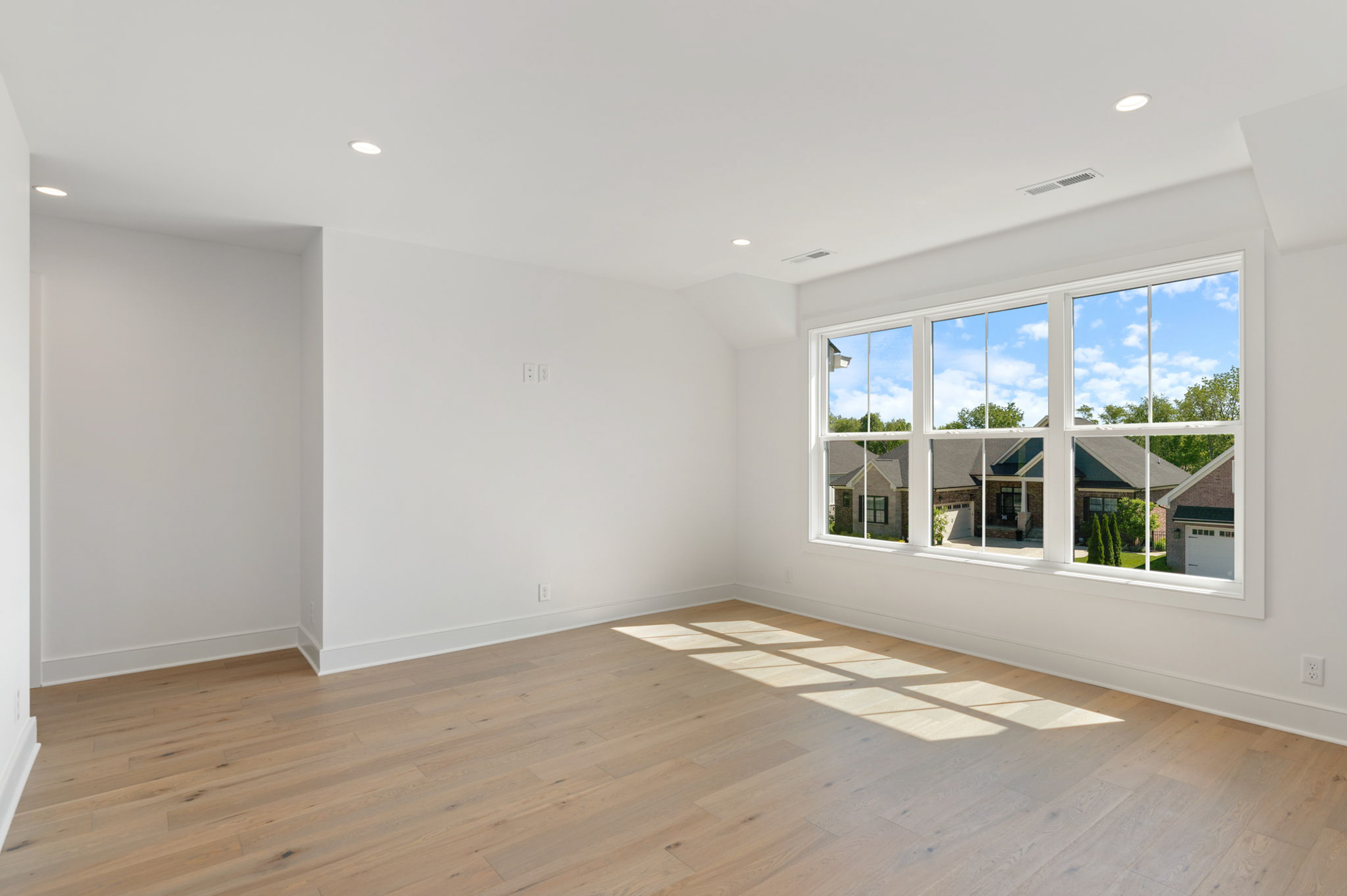 Bright empty living space with a large window and light wood flooring, white walls, and recessed ceiling lights.