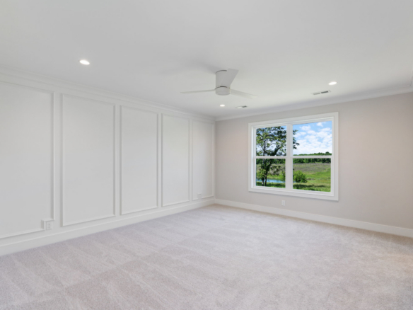Bright empty room with white paneled walls, beige carpet, and a white ceiling fan; window shows trees outside.