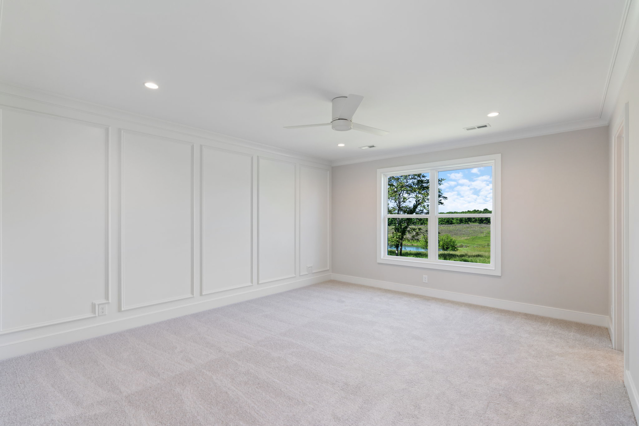Bright empty room with white paneled walls, beige carpet, and a white ceiling fan; window shows trees outside.