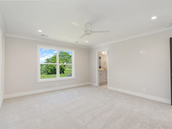Empty bedroom with a white ceiling fan, beige carpet, and light gray walls; large window showing trees outside; doorway to a bathroom visible on the right.