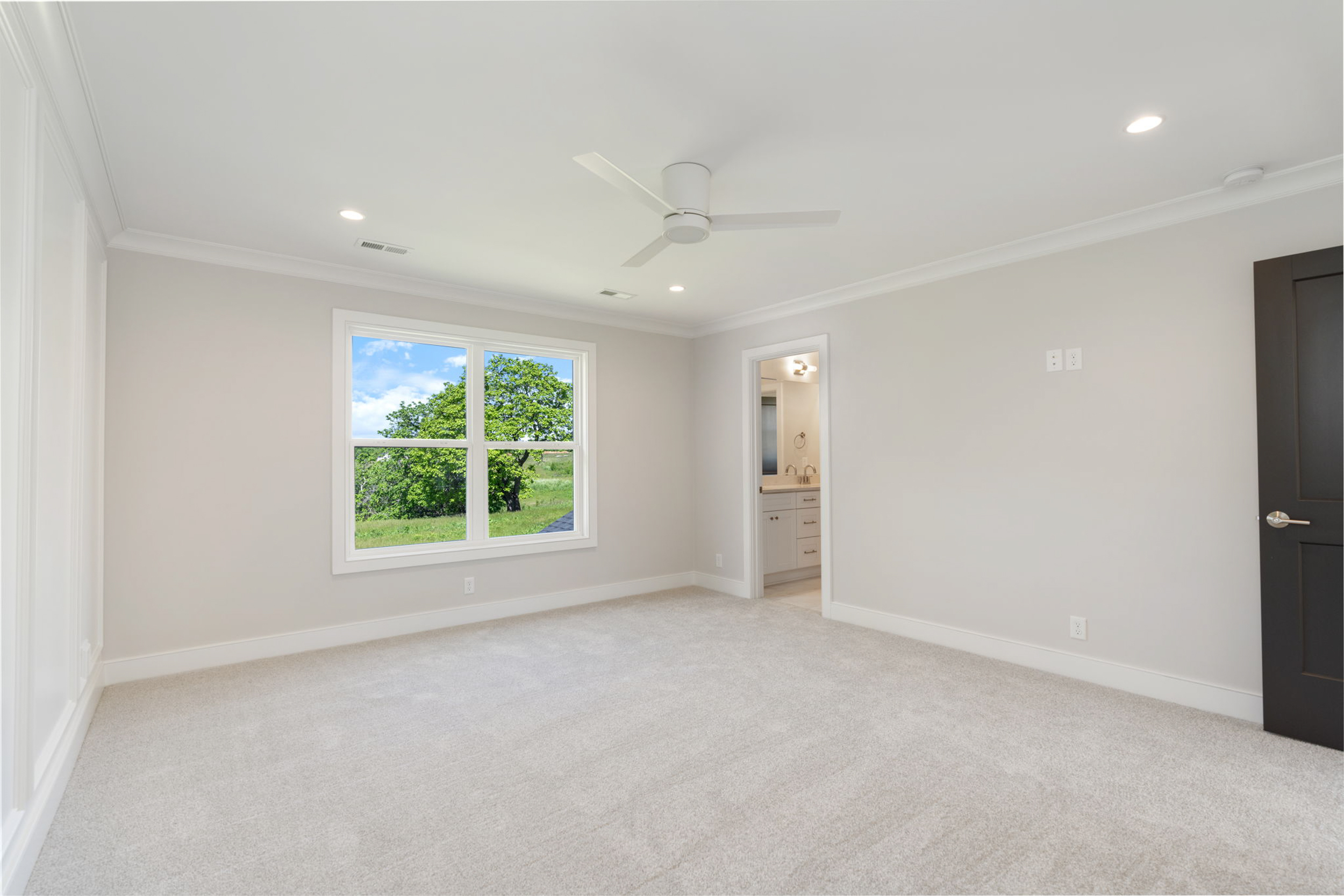 Empty bedroom with a white ceiling fan, beige carpet, and light gray walls; large window showing trees outside; doorway to a bathroom visible on the right.