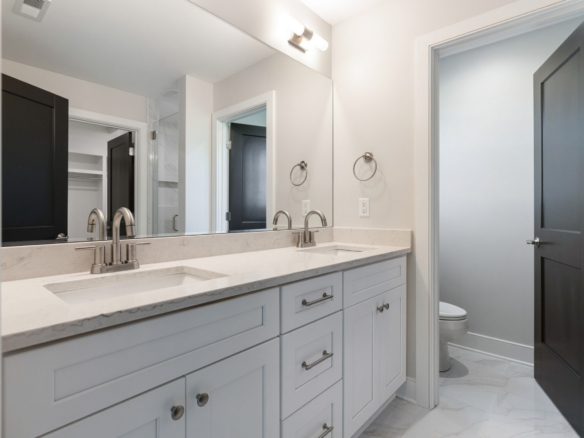 Double-sink bathroom vanity with marble countertop, mirrored wall, and brushed-nickel faucets; open doorway to a separate toilet area with dark doors.