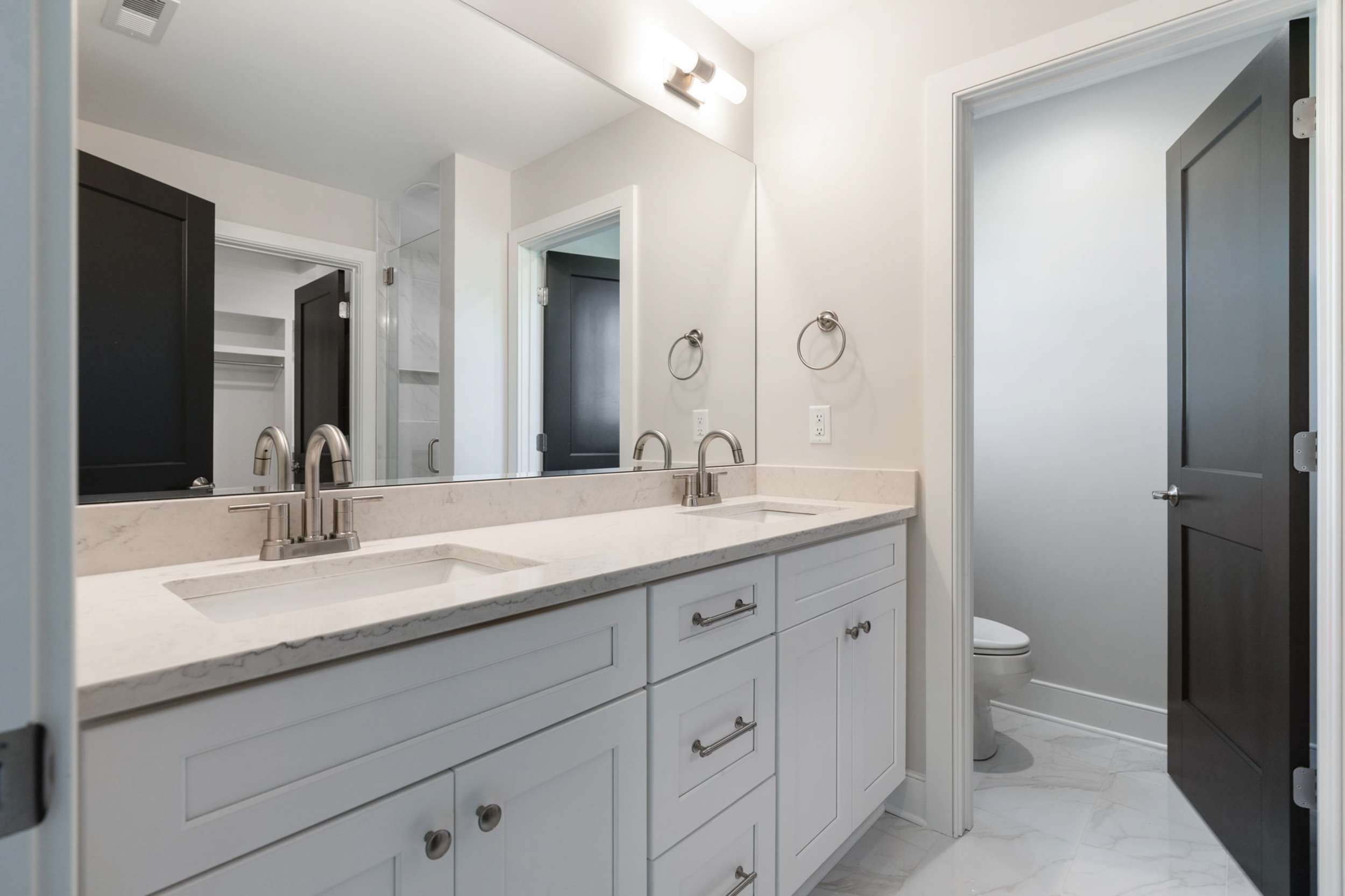 Double-sink bathroom vanity with marble countertop, mirrored wall, and brushed-nickel faucets; open doorway to a separate toilet area with dark doors.