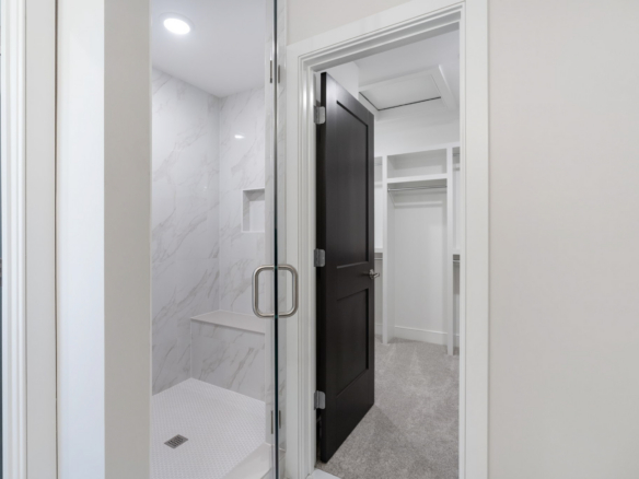 Open bathroom shower with a glass door and chrome handle; white marble tile walls inside; view into a small walk-in closet with white shelving.