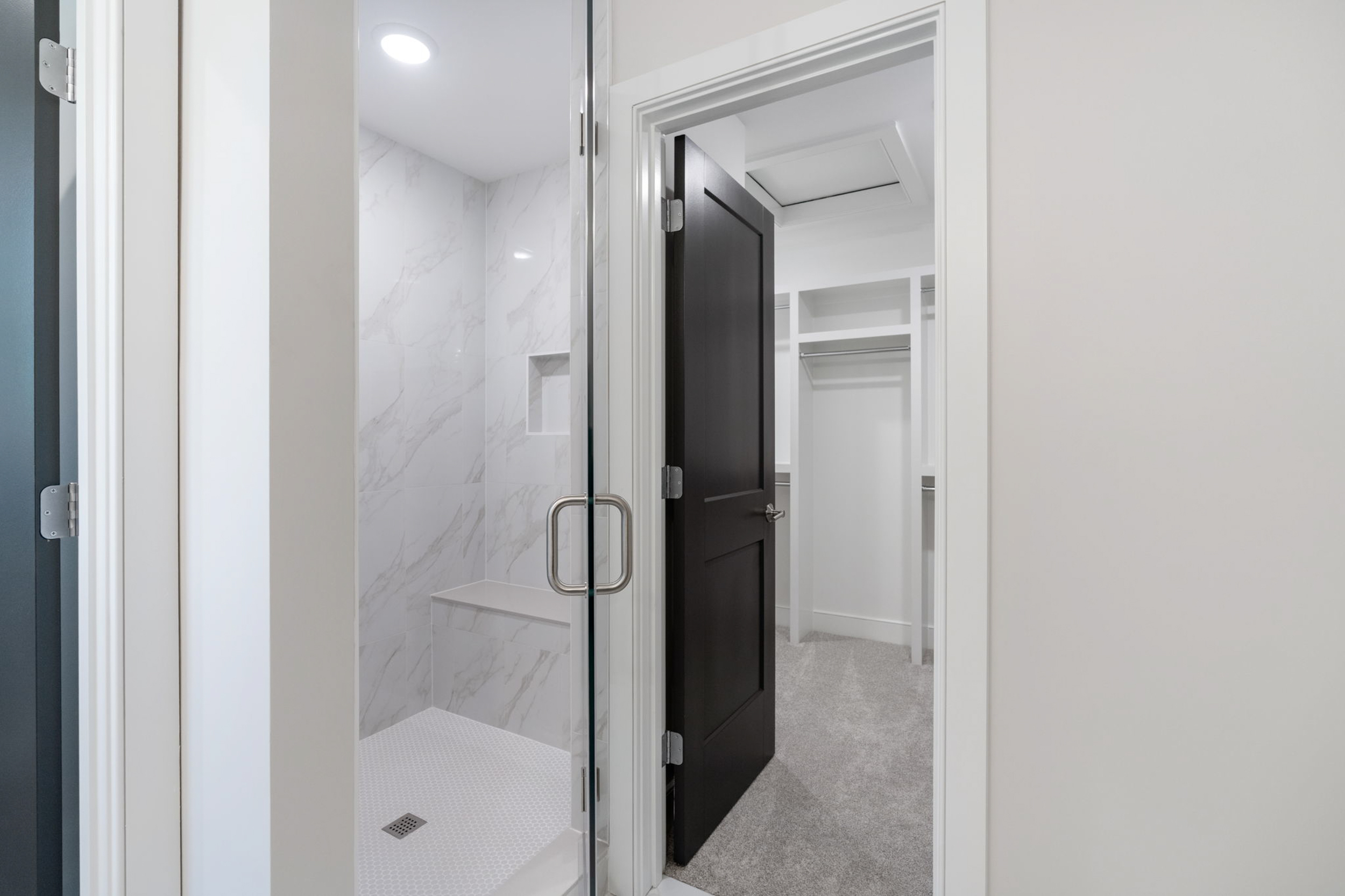 Open bathroom shower with a glass door and chrome handle; white marble tile walls inside; view into a small walk-in closet with white shelving.