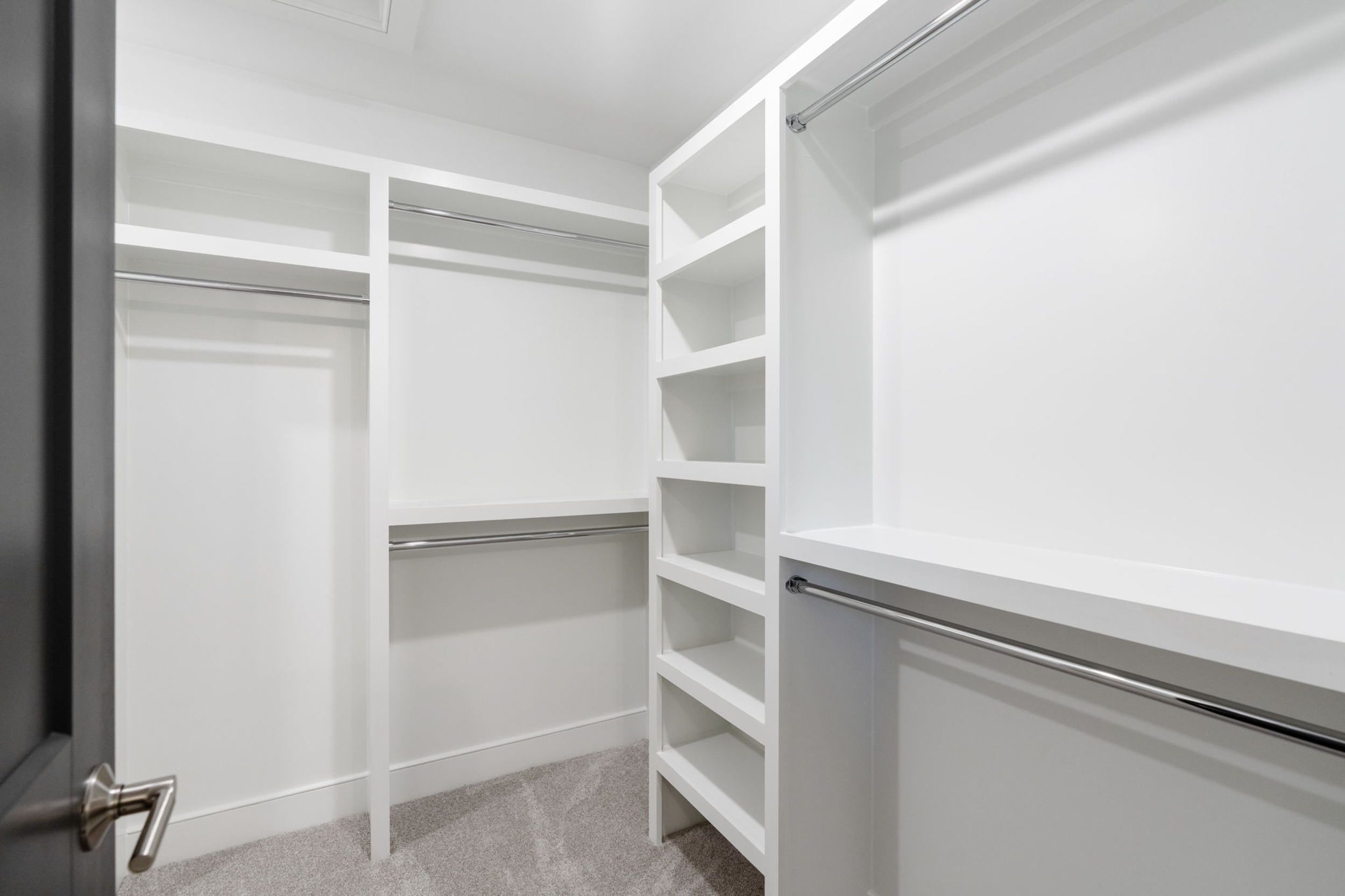 Walk-in white closet with shelving units and double hanging rods along the walls; carpeted floor under bright lighting.