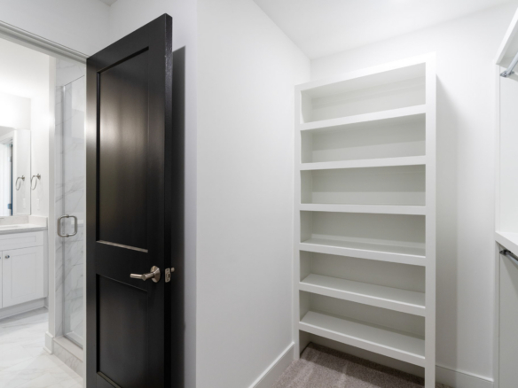 Modern bathroom scene: black door open to a white shelving unit on the right and a glass shower to the left, with marble-tiled floor.