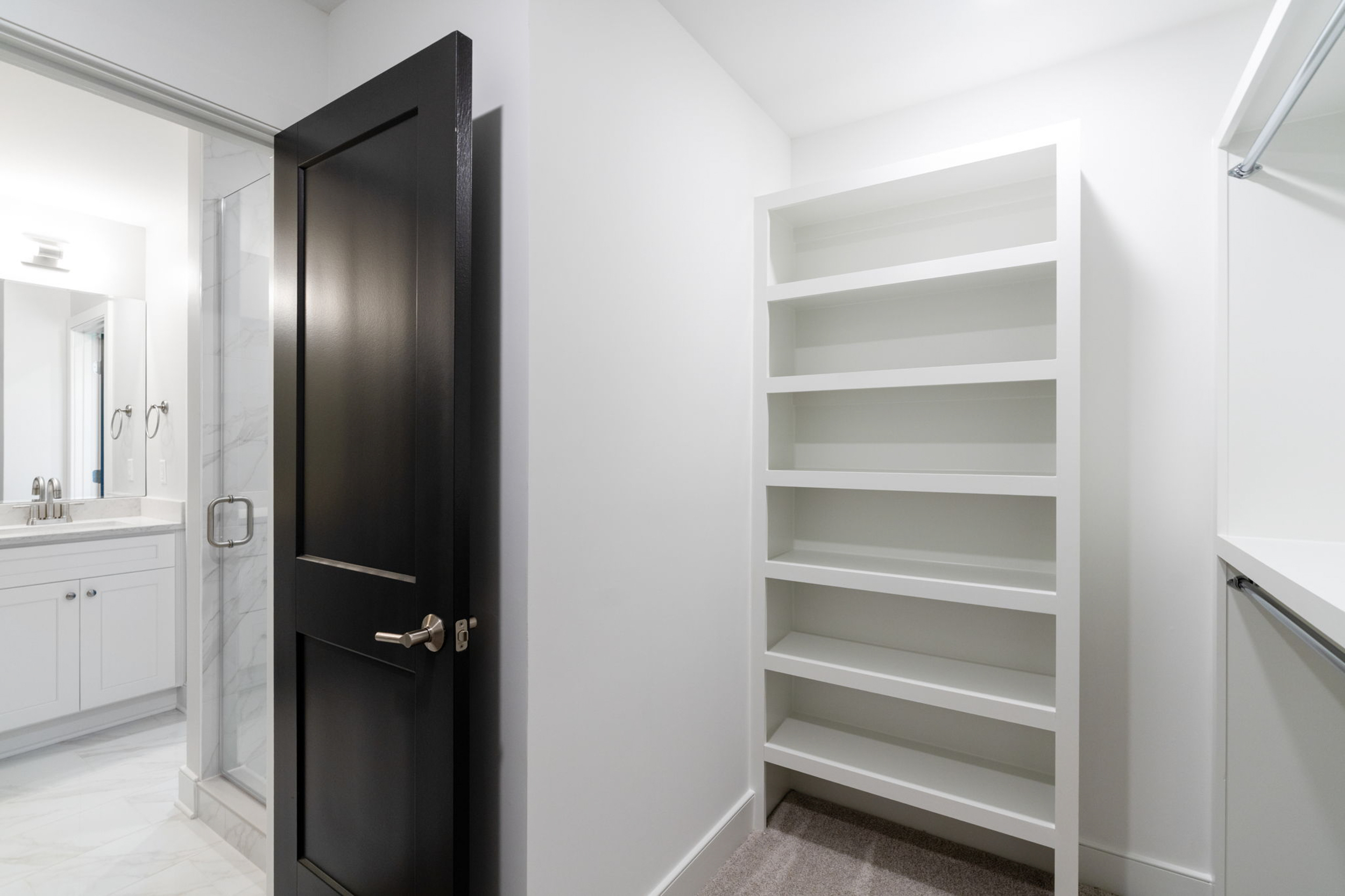Modern bathroom scene: black door open to a white shelving unit on the right and a glass shower to the left, with marble-tiled floor.