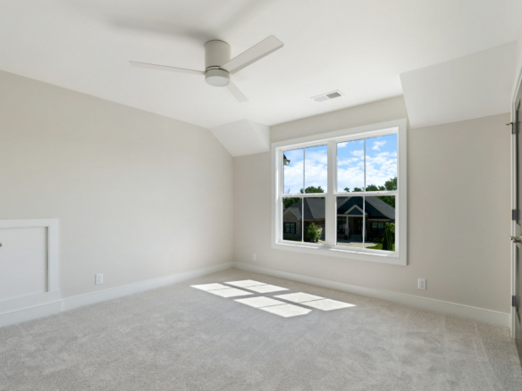 Empty light-filled bedroom with beige walls, white carpet, and a white ceiling fan; a large window reveals a suburban street outside.