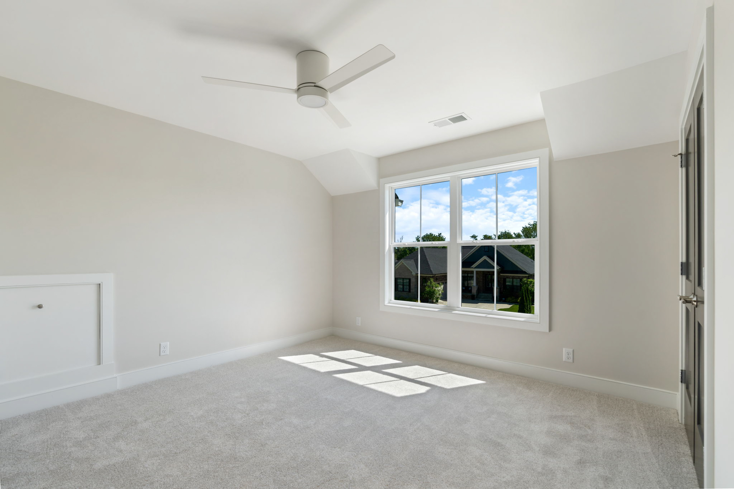 Empty light-filled bedroom with beige walls, white carpet, and a white ceiling fan; a large window reveals a suburban street outside.