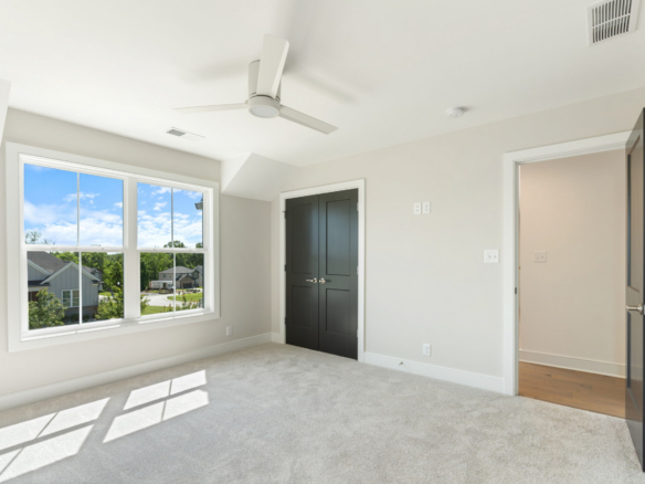 Empty bedroom with gray walls, beige carpet, a large window showing a sunny suburban neighborhood, a white ceiling fan, and dark double doors (open doorway to hall). -or-