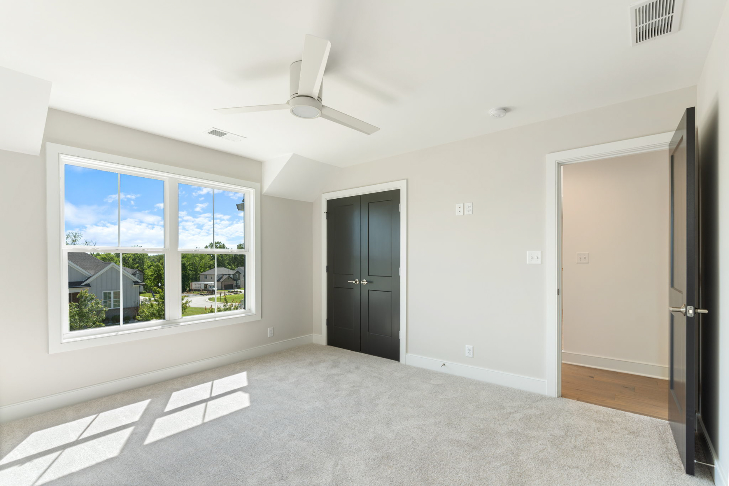 Empty bedroom with gray walls, beige carpet, a large window showing a sunny suburban neighborhood, a white ceiling fan, and dark double doors (open doorway to hall). -or-