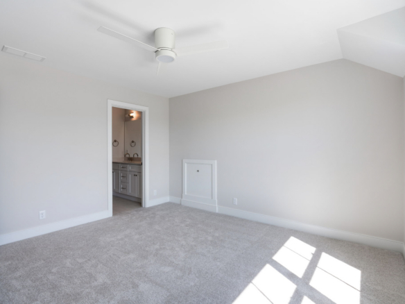 Empty modern bedroom with white ceiling fan, light gray walls, beige carpet, and doorway to an adjacent bathroom; sunlight casts through the window on the right.