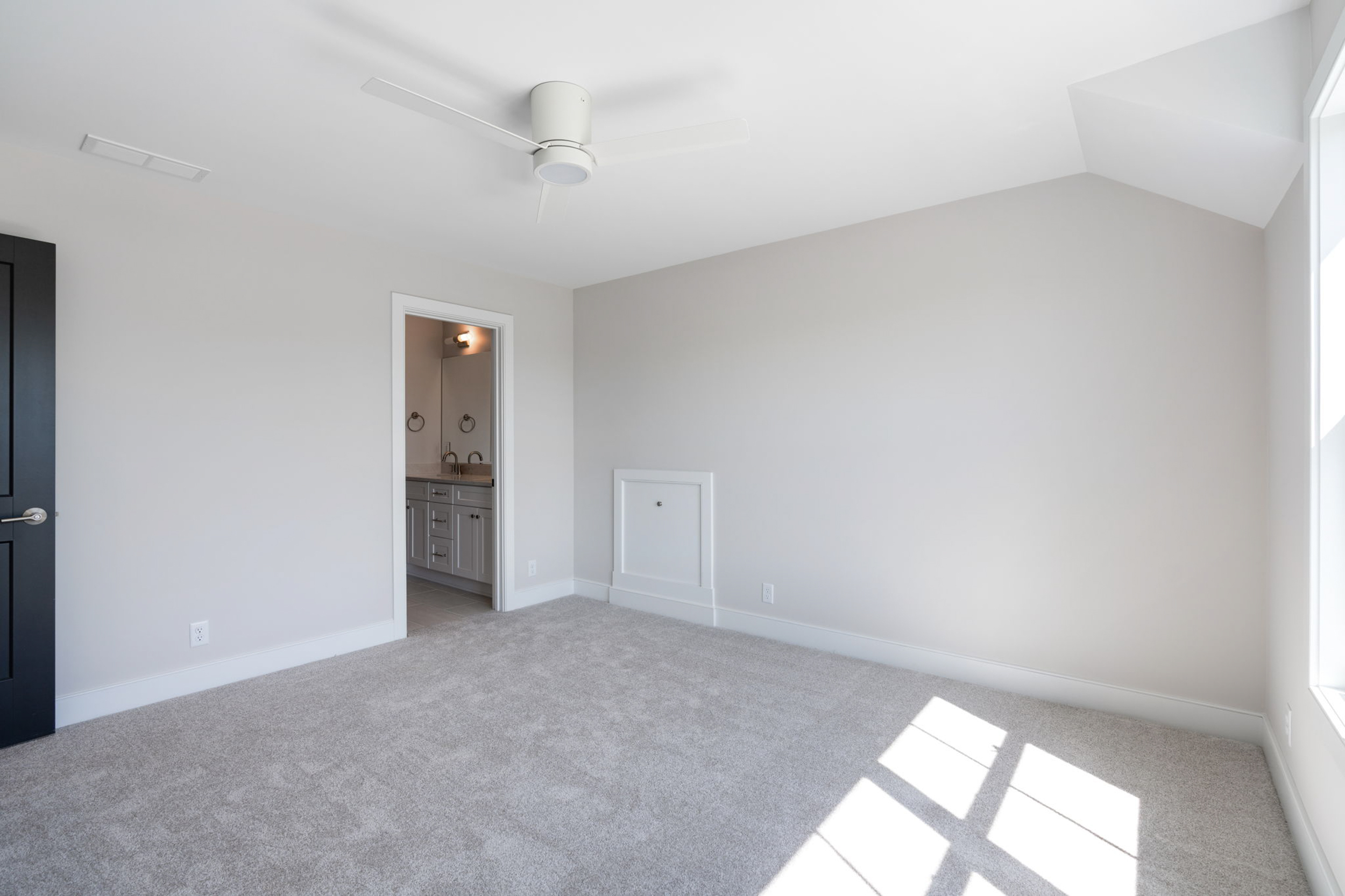 Empty modern bedroom with white ceiling fan, light gray walls, beige carpet, and doorway to an adjacent bathroom; sunlight casts through the window on the right.