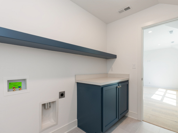 Laundry utility area with dark blue cabinets, light countertop, and a blue wall shelf; washer hookups on wall and doorway to adjacent bright room.