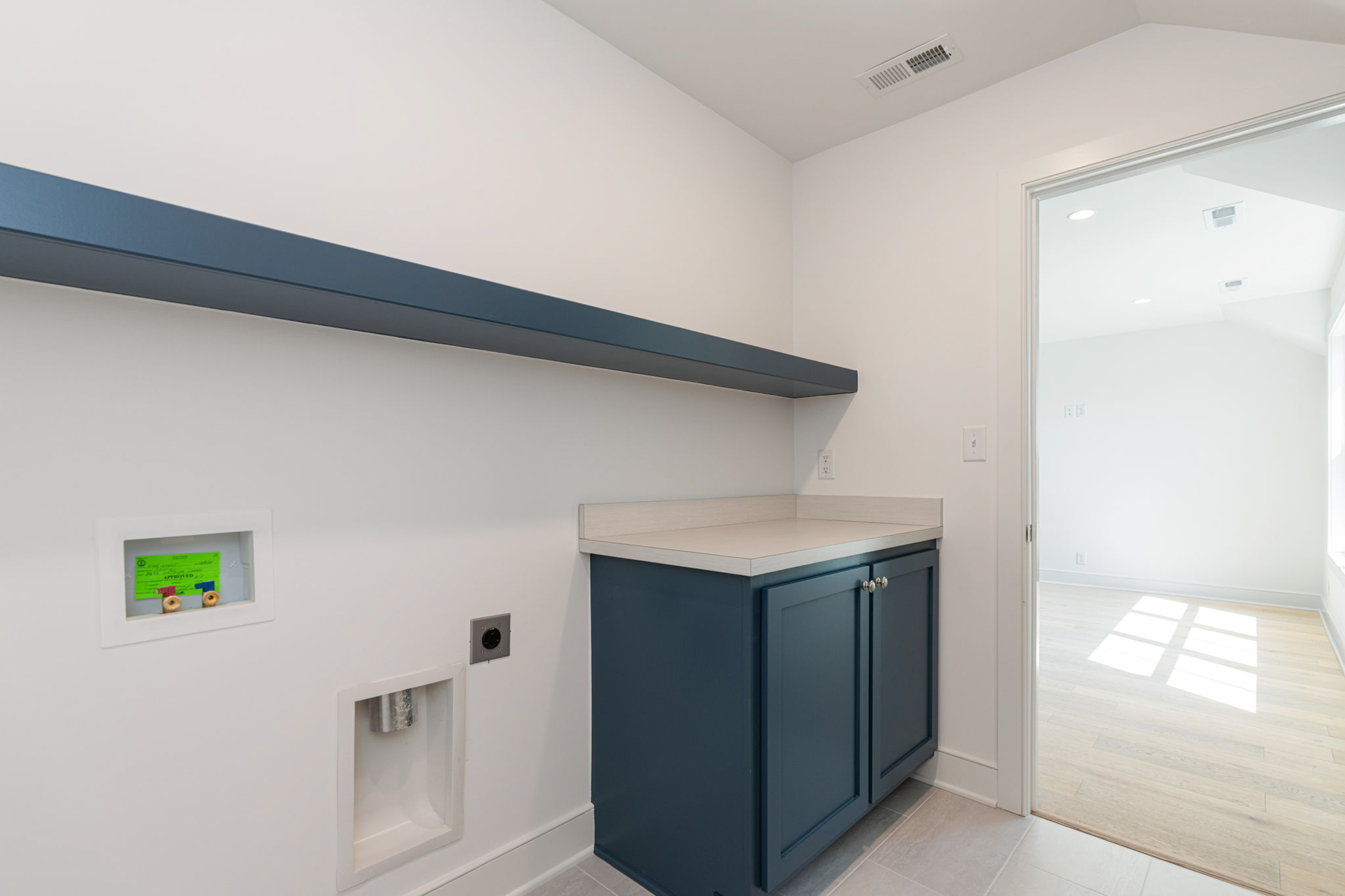 Laundry utility area with dark blue cabinets, light countertop, and a blue wall shelf; washer hookups on wall and doorway to adjacent bright room.