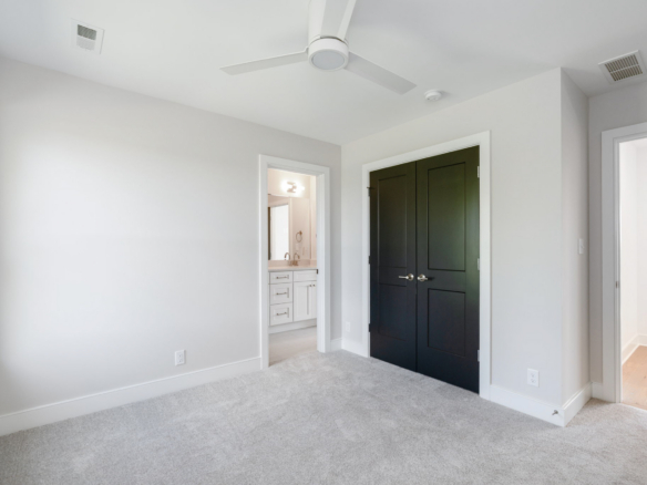 Bright bedroom with light gray walls, white trim, and a white ceiling fan; a pair of dark double doors stands opposite. A bathroom doorway with a vanity is visible.