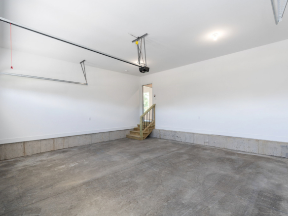 Empty residential garage with concrete floor, white walls, overhead door track, and a wooden staircase at the back corner.