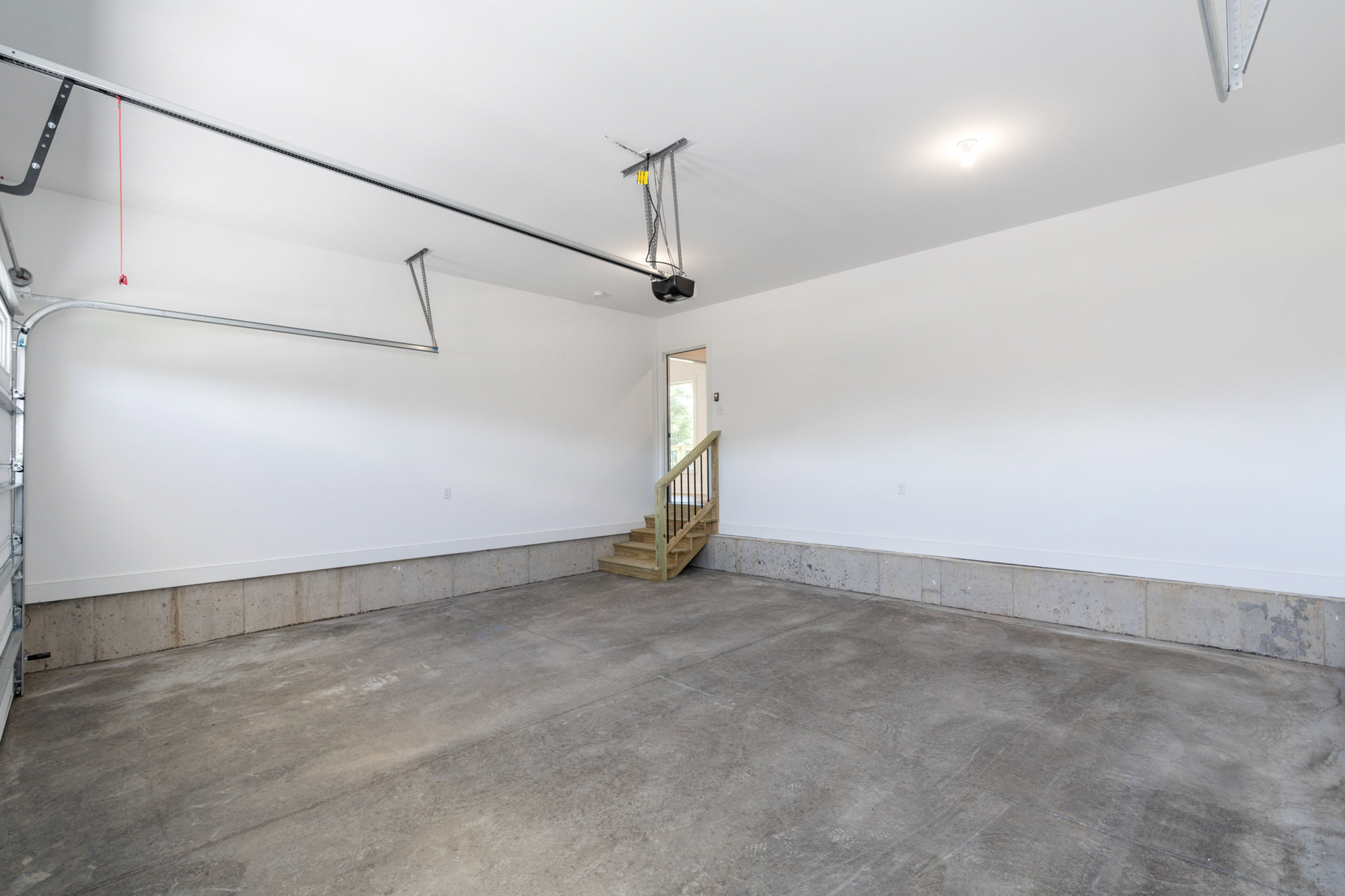 Empty residential garage with concrete floor, white walls, overhead door track, and a wooden staircase at the back corner.