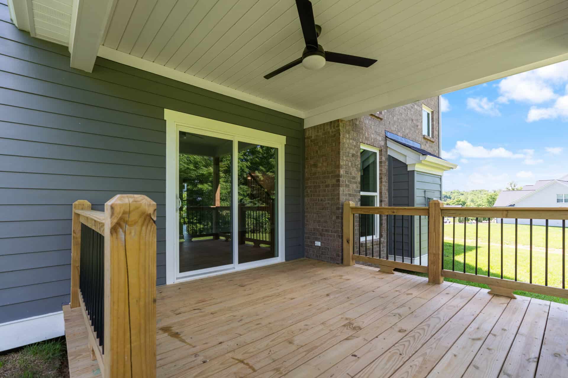 Wooden back deck attached to a gray house with sliding glass doors, a ceiling fan, and a wooden railing overlooking a yard.