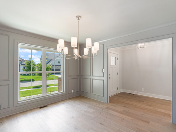 Empty dining area with a modern chandelier, gray paneled walls, and light wood flooring; large window shows a sunny suburban street.