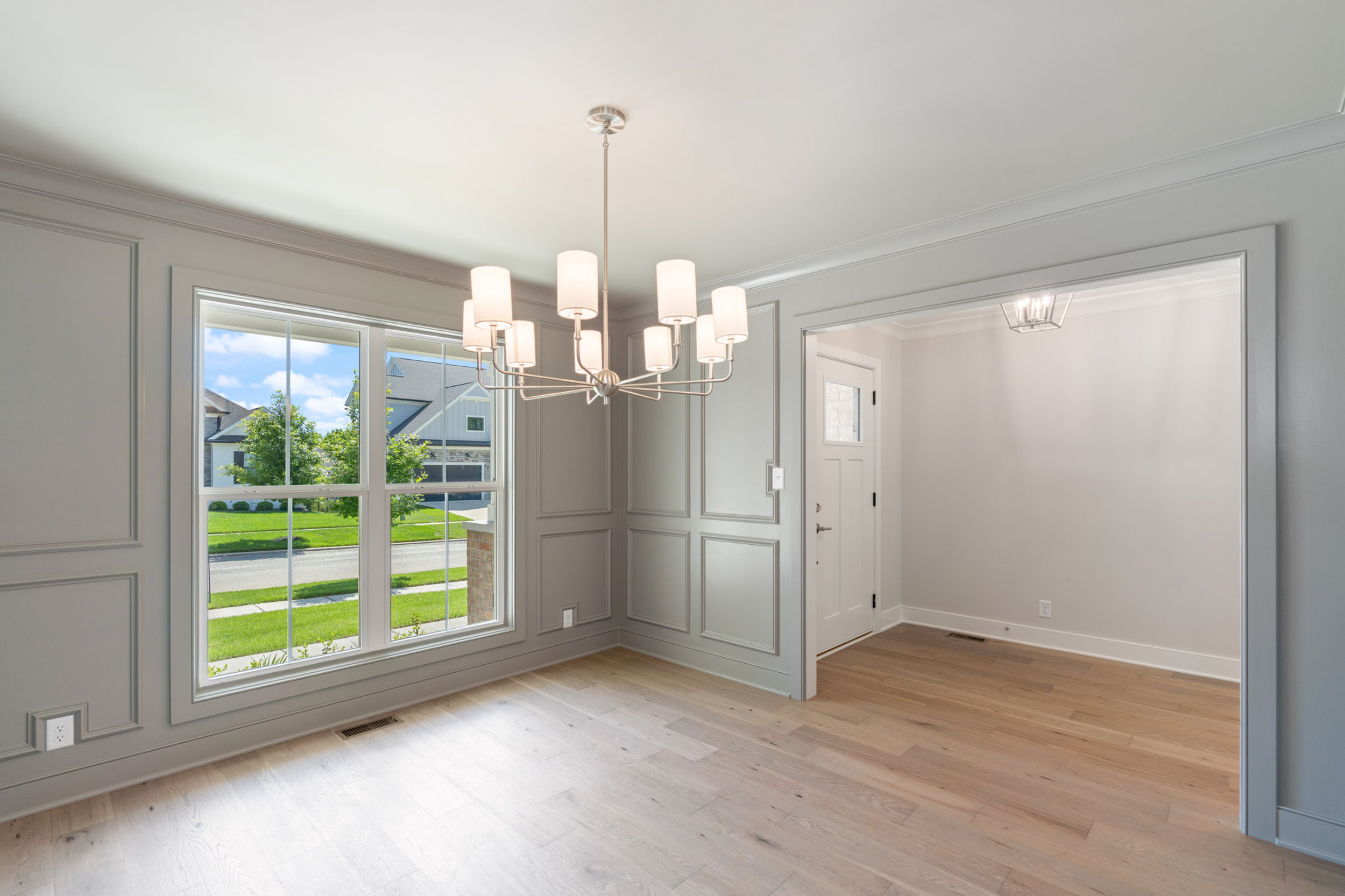 Empty dining area with a modern chandelier, gray paneled walls, and light wood flooring; large window shows a sunny suburban street.
