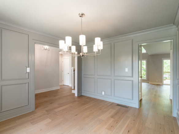 Hallway with gray paneled walls, a metal chandelier, and light hardwood flooring; open doorways to adjacent rooms and a sunny window view.