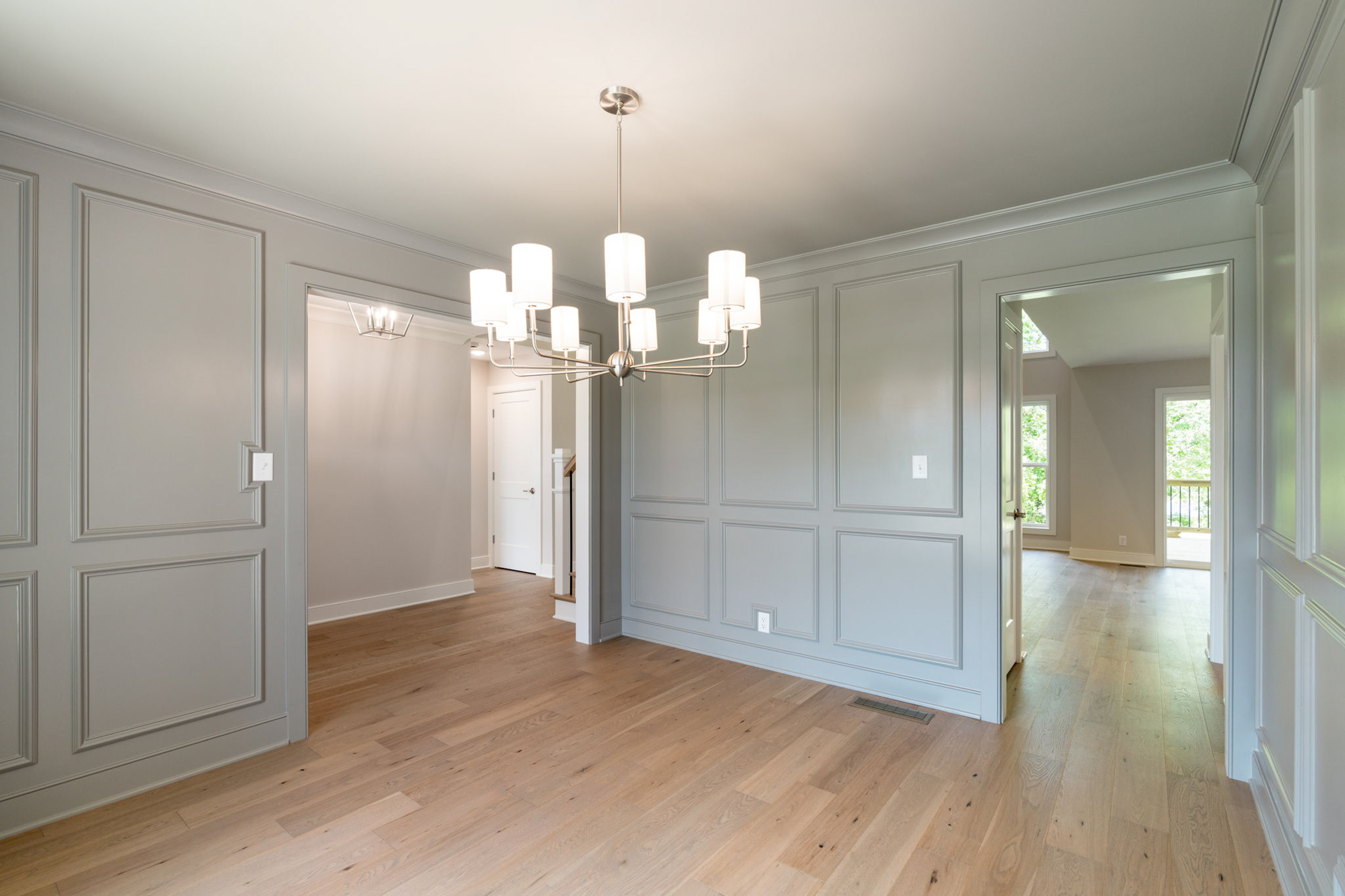 Hallway with gray paneled walls, a metal chandelier, and light hardwood flooring; open doorways to adjacent rooms and a sunny window view.