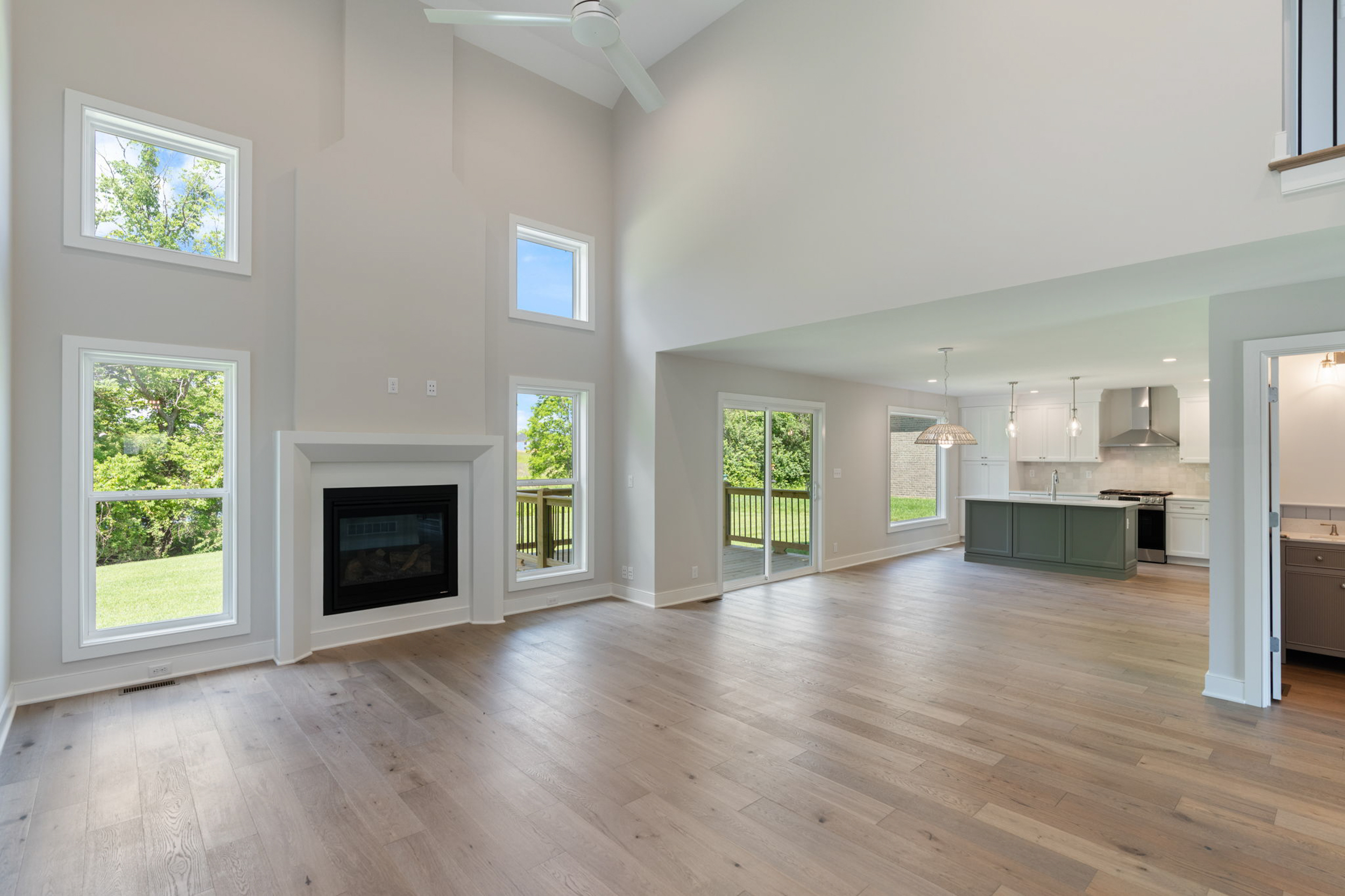 Open living area with vaulted ceiling, large windows, a white-framed fireplace, and a kitchen island in the background. (Open-concept space)