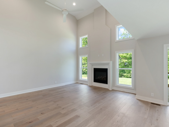 Bright living room with light wood floors, a white fireplace, and tall vaulted ceiling with windows showing greenery outside