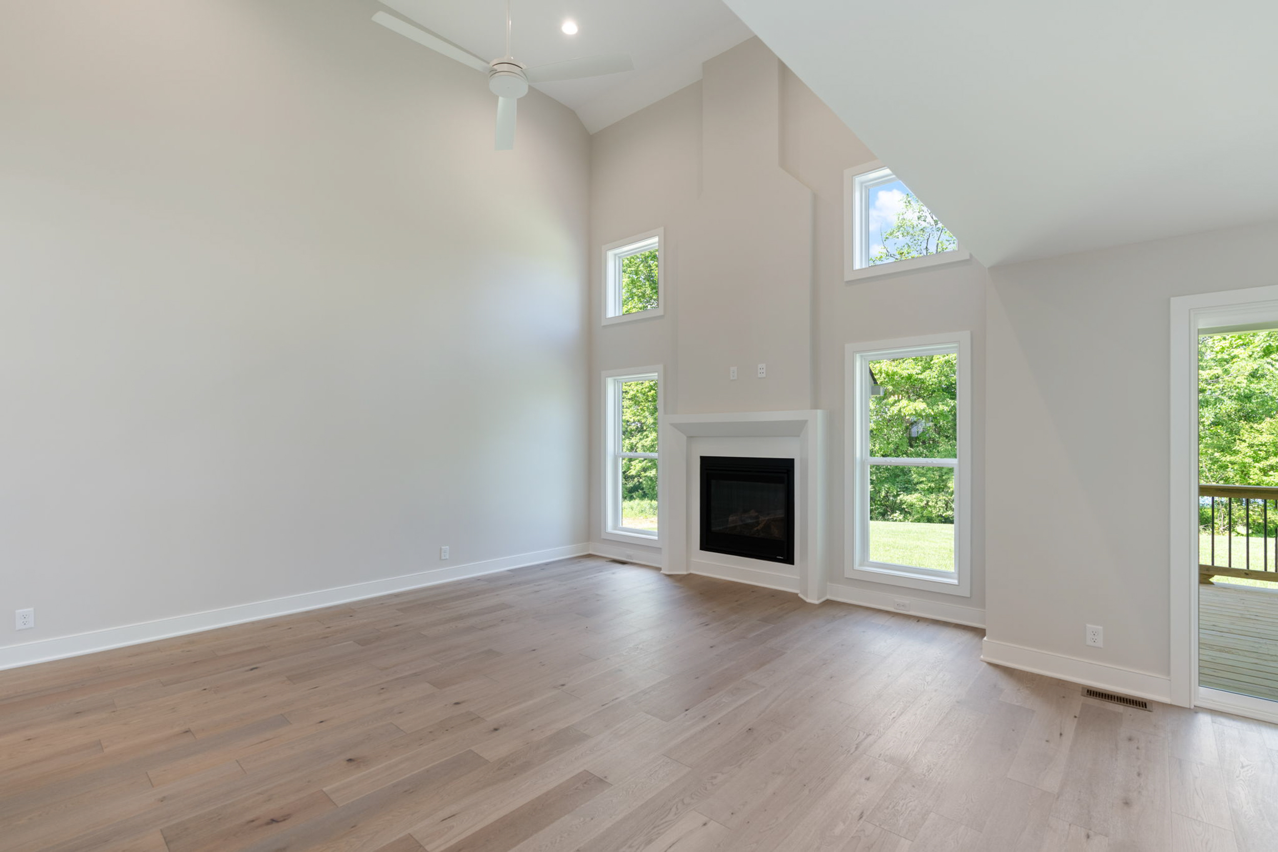 Bright living room with light wood floors, a white fireplace, and tall vaulted ceiling with windows showing greenery outside