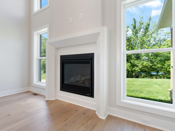 Bright living room with a white fireplace surround and large windows showing a green, sunny yard outside.",