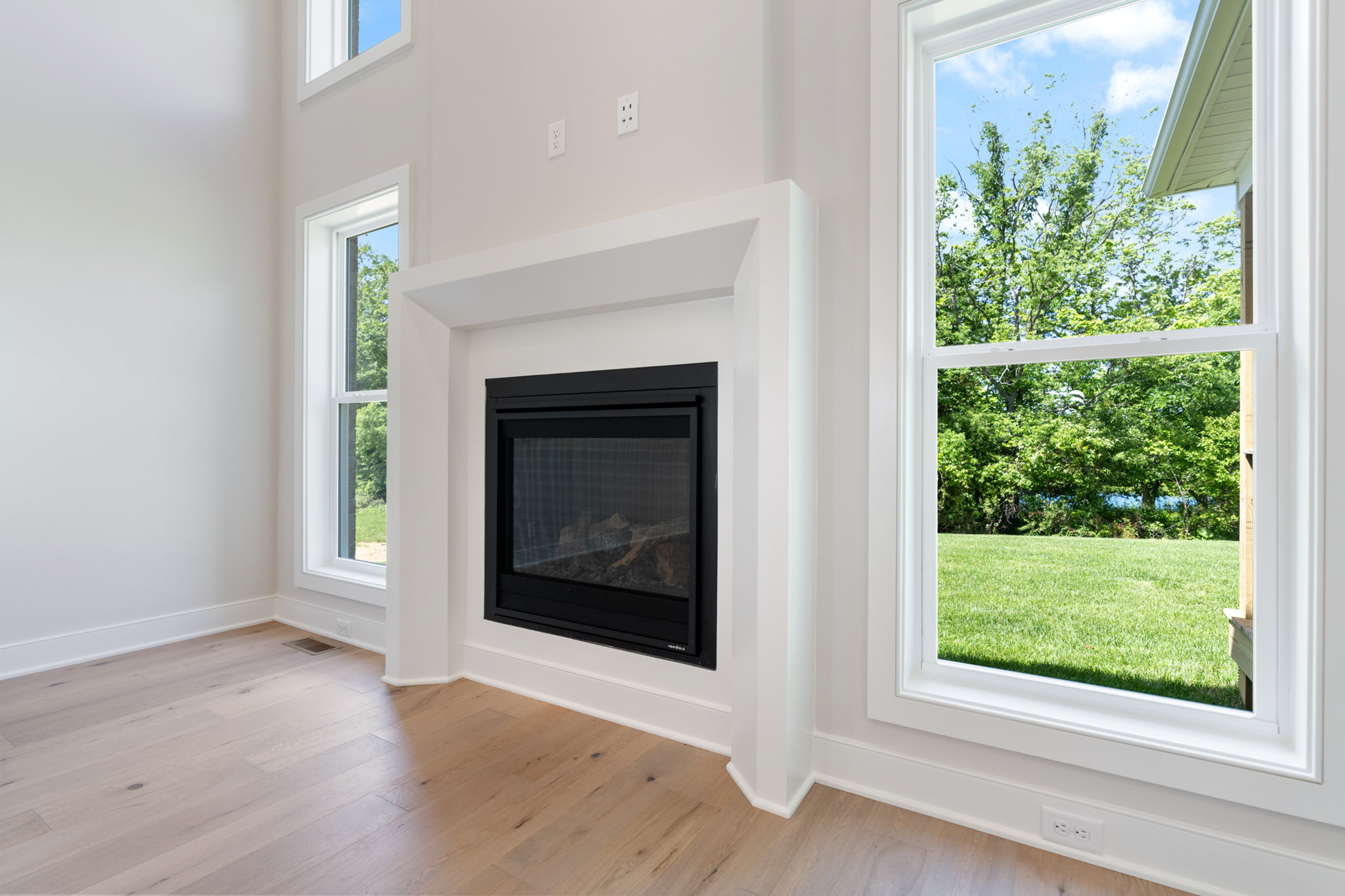 Bright living room with a white fireplace surround and large windows showing a green, sunny yard outside.",