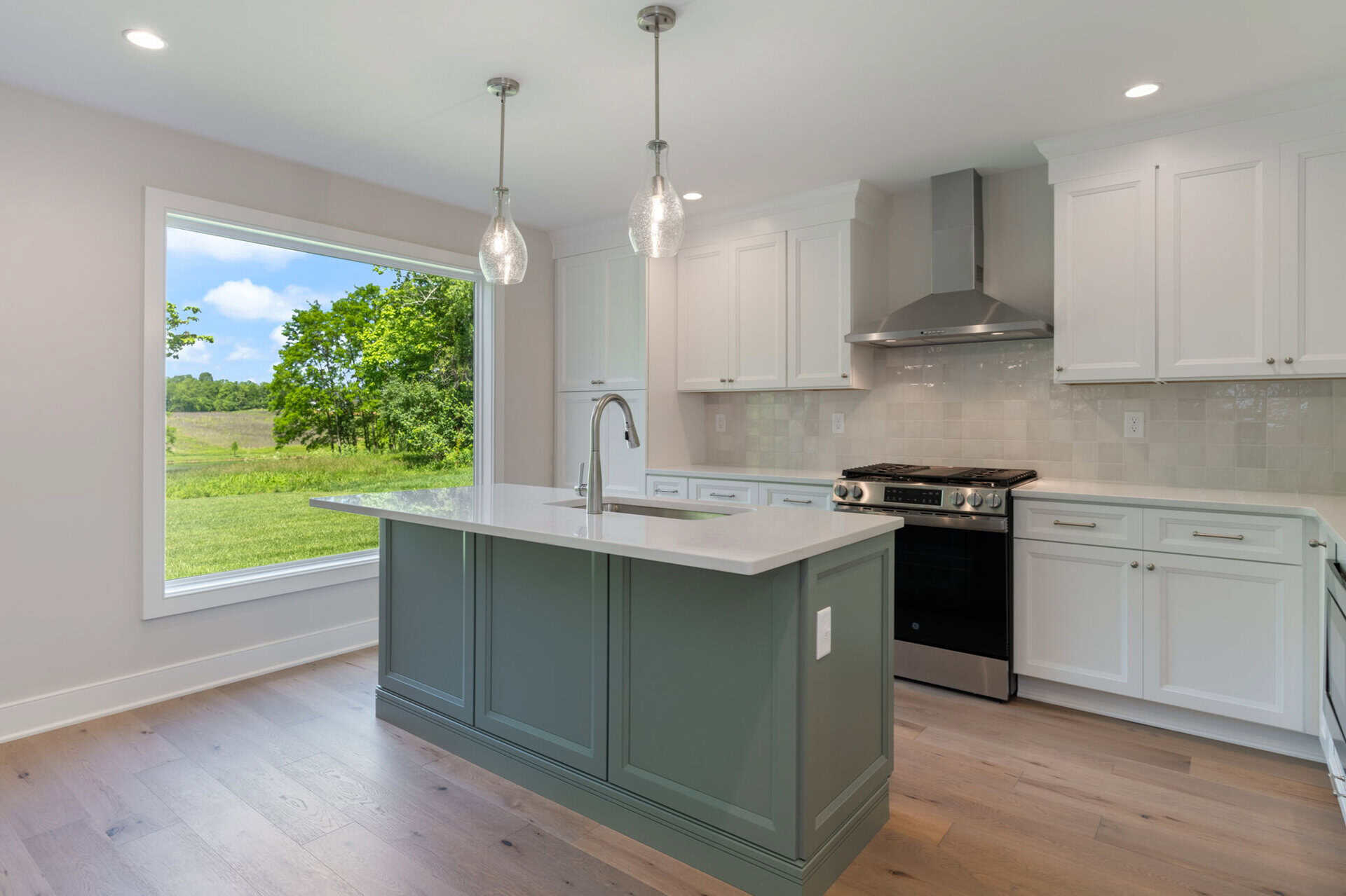 Modern kitchen with white cabinets, gray island, and large window overlooking green landscape.