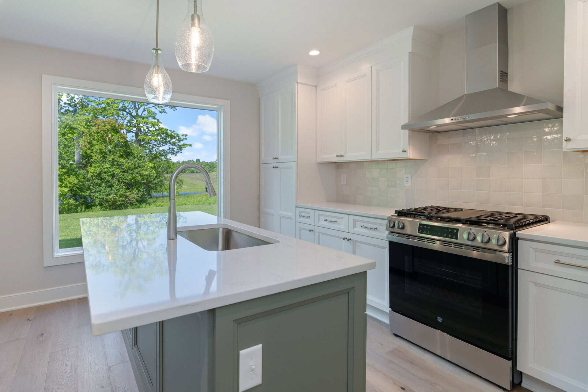 Modern kitchen with white cabinets, stainless steel stove, and large window showing greenery.