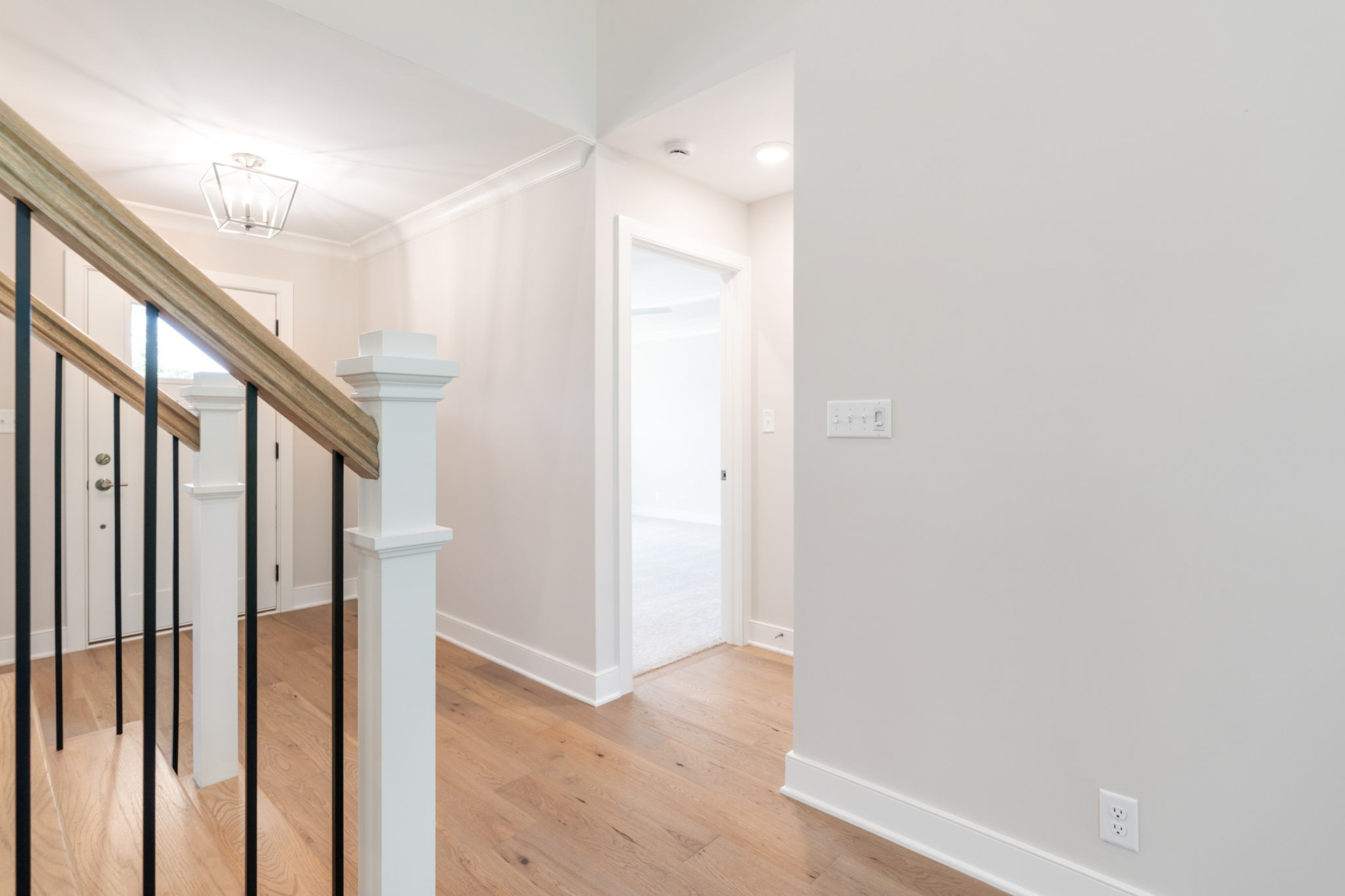 Interior hallway with a wooden staircase and black metal balusters, bright lighting, and light gray walls.