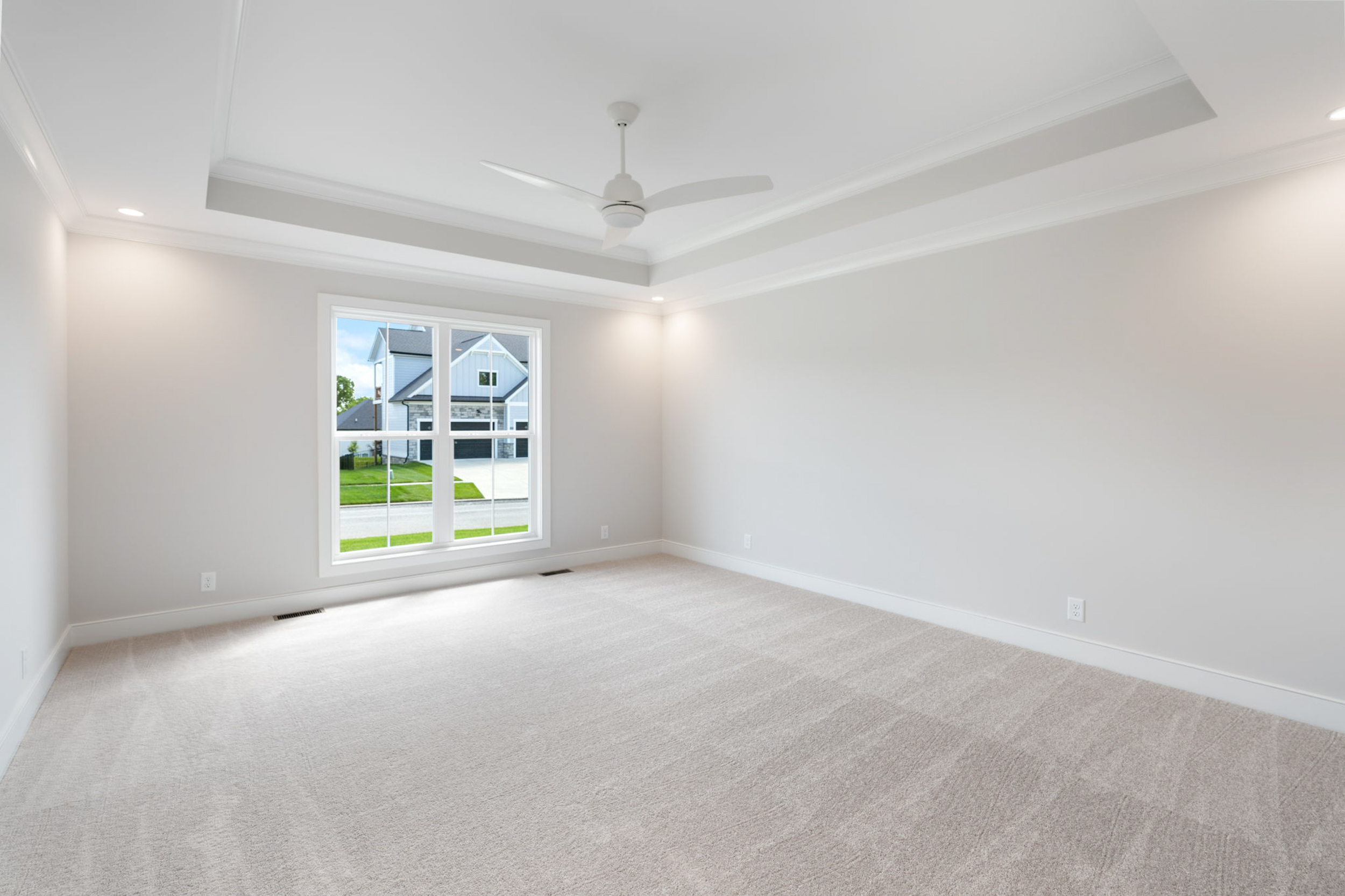Bright empty bedroom with beige carpet, white ceiling fan, and a large window letting in natural light from outside the home.] ,