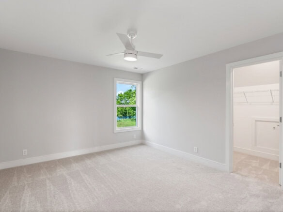 Empty bedroom with white walls, carpet, ceiling fan, and window showing greenery outside.