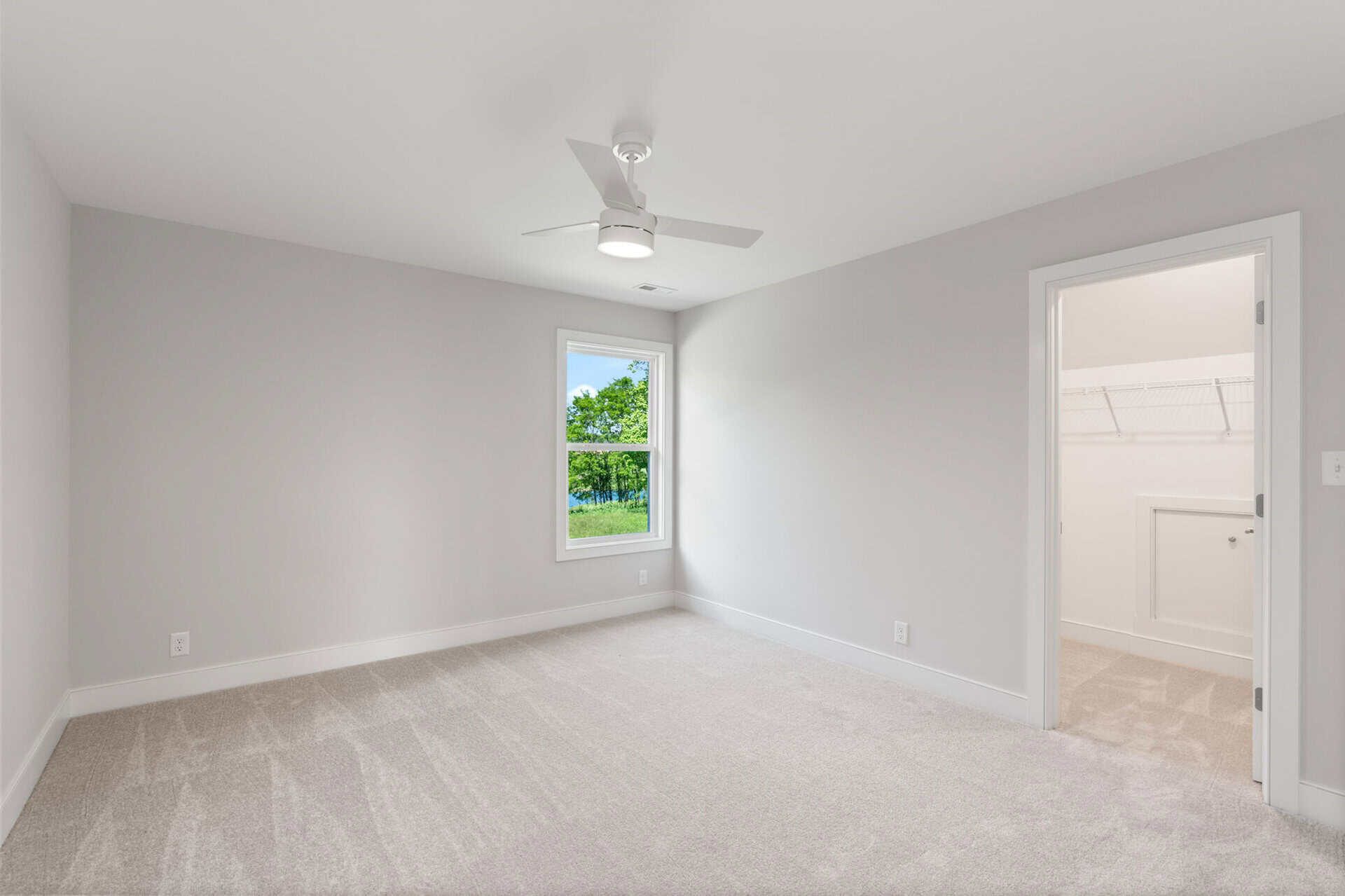 Empty bedroom with white walls, carpet, ceiling fan, and window showing greenery outside.