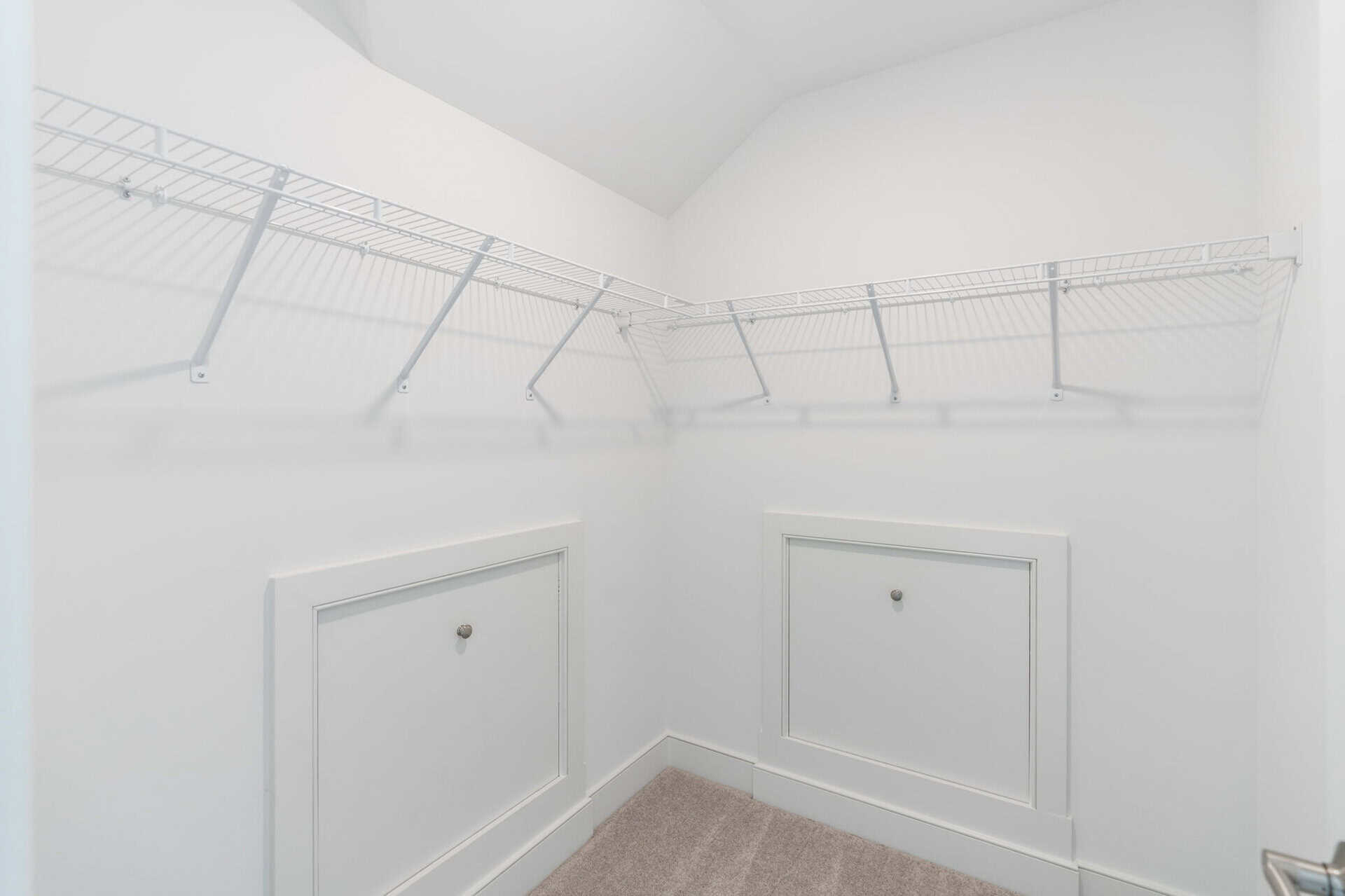 White walk-in closet with metal wire shelving along two walls and built-in lower cabinets; light beige carpet on floor.