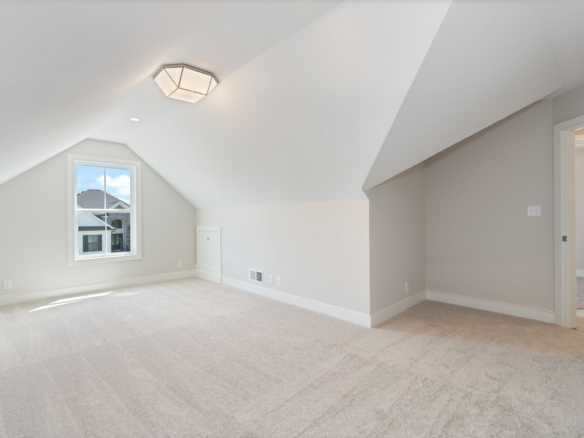 Empty attic bedroom with sloped white ceilings, beige carpet, and a window showing houses outside.